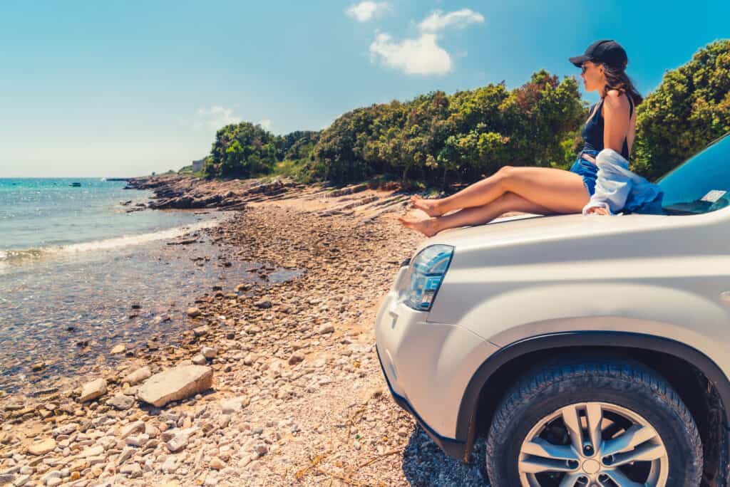 Buying a car in Australia A woman wearing a black cap and sunglasses sits on the hood of a white SUV parked on a pebbly shoreline, with her legs stretched out, facing the sea. Trees line the coast under a clear blue sky.